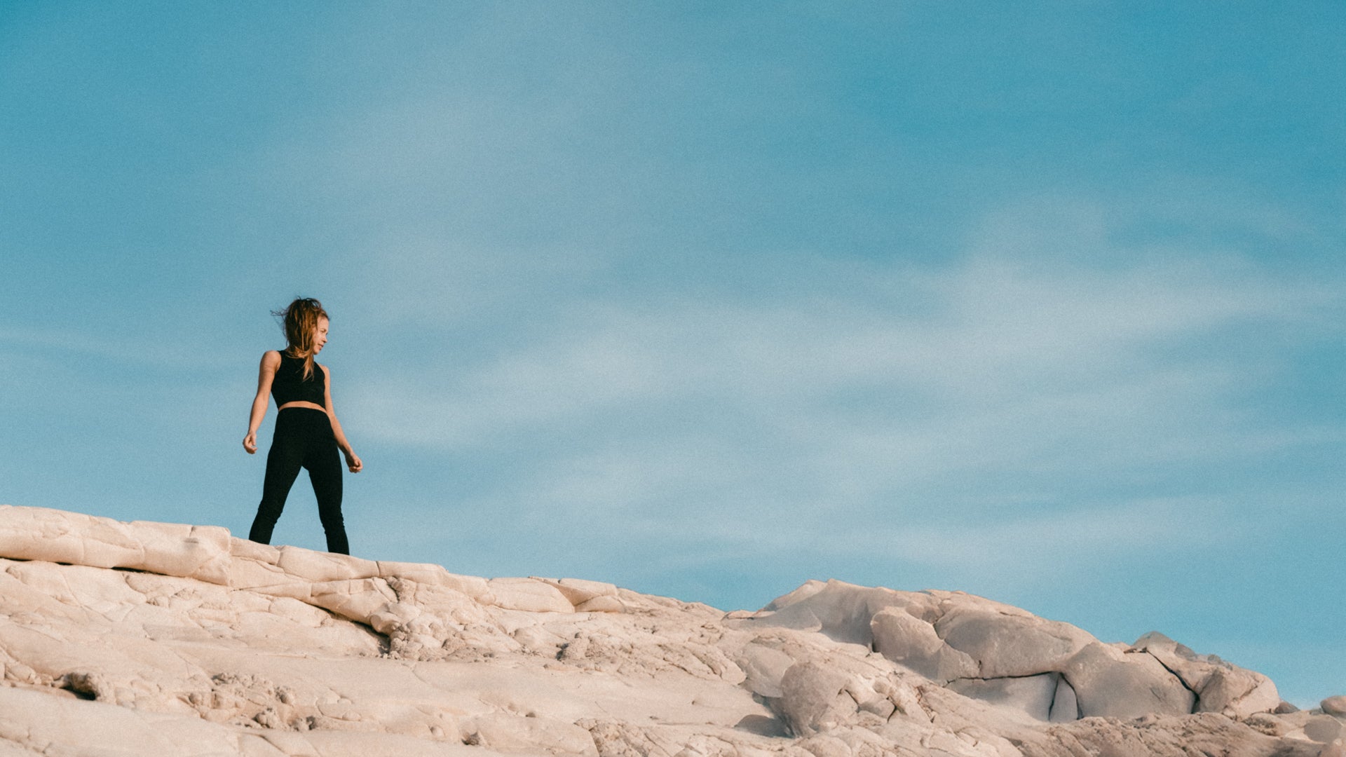  Woman in minimalist sustainable luxury activewear set exploring white coastal rocks against turquoise sky - Ainé hybrid top and leggings by Studio Nidra