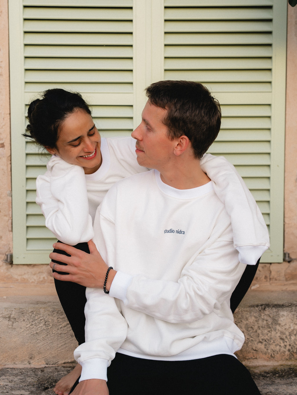 Two people in The Crew Sweatshirt limited edition in Marshmallow White sitting together in front of a building with green shutters.